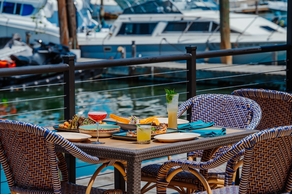 People enjoy food and drinks at a table by water with boats in the background.